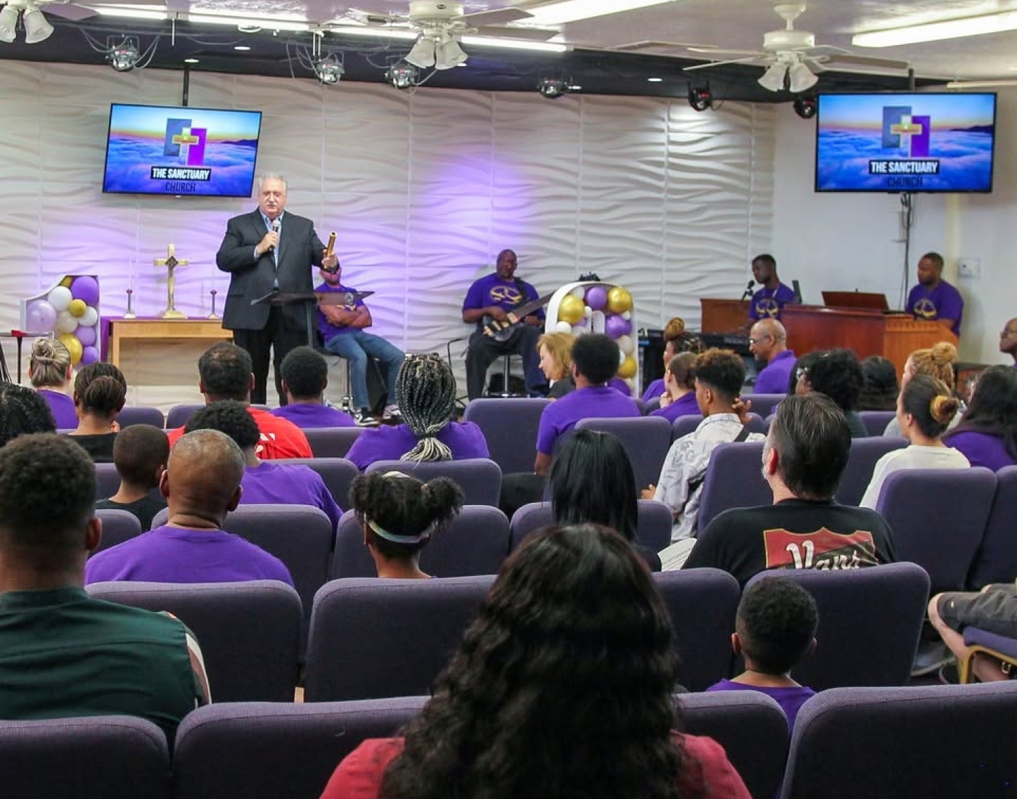 Congregation gathered inside The Sanctuary Church during a worship service with a speaker and musicians at the front.