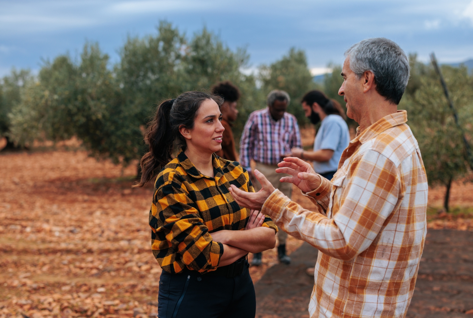 Two people discussing agricultural work outdoors, with other workers in the background.